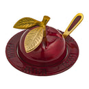 A deep red, apple-shaped honey pot with a golden leaf and stem detail, resting on a matching textured red saucer, with a small golden spoon emerging from the back.