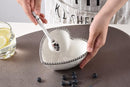 A white heart-shaped porcelain bowl with a silver beaded rim, accompanied by a matching white spoon. Dimensions of the bowl (5.5" x 5.5" x 2.5") and spoon (5" long) are indicated.