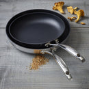 Two stacked ScanPan HaptIQ fry pans on a wooden surface with scattered grains