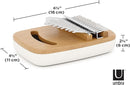 A slightly elevated side-angle view of an Umbra Strumba Kalimba, a small musical instrument. It has a light wooden top surface featuring a crescent-shaped sound hole and a set of metallic tines secured by a silver-toned bridge. The base of the instrument is a smooth, rounded, off-white or cream color. The image is set against a plain white background. The brand logo "U umbra" is visible in the bottom right corner. Additionally, dimension lines with measurements "6 1/2" (16 cm)" for length and "2 1/2" (6 cm)