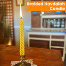 Yellow braided Havdalah candle burning on a table in a bright, cozy indoor space labeled “Braided Havdalah Candle”