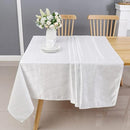A white tablecloth drapes over a wooden table, featuring a vase of flowers at its center, with two wooden chairs in the background.