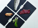 The image shows a Joseph Joseph Folio 4-Piece Cutting Board Set. It consists of a sleek, dark-colored (likely black or dark gray) storage case that holds four individual cutting boards. The tops of the cutting boards are visible, showing different colored tabs, which are typically used for hygienic separation of food types (e.g., raw meat, vegetables, cooked food, fish). The Joseph Joseph logo is visible on the lower front of the case.