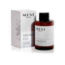 A 100ml bottle of SCENT New York Aroma Oil in the 'Paradise' fragrance. The amber glass bottle has a minimalist white label with black text and a black cap. Next to the bottle is its matching packaging box, which has a similar white and black design with subtle floral artwork on the side.