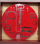 A striking red circular wall clock with large, stylized black numerals, featuring a clear vertical stripe through the center with the brand "HOME" subtly visible. The clock is shown inside an open brown cardboard box.