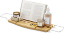 A light wood Umbra bathtub caddy with expandable metal arms, featuring a slotted book/tablet stand, a wine glass holder, and a recessed area with drain holes for soap or accessories.