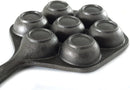 The image displays a cast iron aebleskiver pan. It's a dark-colored pan with a single handle and seven hemispherical indentations, which are used to cook aebleskiver (Danish pancake balls). The shape of the pan, excluding the handle, is hexagonal.