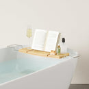 A light wood Umbra bathtub caddy with expandable metal arms, featuring a slotted book/tablet stand, a wine glass holder, and a recessed area with drain holes for soap or accessories.