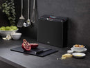 The image shows a Joseph Joseph Folio 4-Piece Cutting Board Set. It consists of a sleek, dark-colored (likely black or dark gray) storage case that holds four individual cutting boards. The tops of the cutting boards are visible, showing different colored tabs, which are typically used for hygienic separation of food types (e.g., raw meat, vegetables, cooked food, fish). The Joseph Joseph logo is visible on the lower front of the case.
