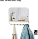 A sleek white wall-mounted organizer mirror with rounded corners, featuring a light wood shelf beneath it. Below the shelf, a white bar extends with several wooden peg hooks.