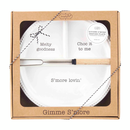 A Mud Pie Circa S'mores Divided Serving Plate and Skewer Set presented in a brown cardboard gift box with a clear window. The white ceramic plate has three sections and beaded edges. One section has "Melty goodness" printed on it, another has "Choc it to me," and the largest section says "S'more lovin'". A metal skewer with a wooden handle rests across the plate. The box is tied with black and white twine and has a label that reads "DIVIDED S'MORE SERVING PLATE & ROASTING FORK SET". The words "Gimme S'more"