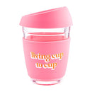 Clear glass travel mug with a bright pink silicone sleeve and lid, adorned with the phrase "living cup to cup," by Mud Pie.
