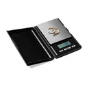 High-precision black digital pocket scale with a stainless steel weighing platform. Two gold rings, one with a small stone, are placed on the scale, which displays a reading of "3.9". The scale has a hinged black cover that is partially open.