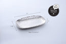 A Pampa Bay Millennium Titanium-Plated Porcelain 11-inch Platter is displayed on a white background. The silver-colored platter features a textured, hammered finish and slightly raised, irregular edges. Dimensions are shown: 11" length, 7.25" width, 1" height. A speech bubble reads, "I CAN'T BELIEVE IT'S NOT METAL! IT'S PORCELAIN," and lists features: freezer/dishwasher safe, tarnish/stain/scratch resistant. "PAMPA BAY" is also included.