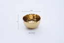 A slightly angled view of a Pampa Bay Golden Millennium Porcelain Snack Bowl. The bowl has a rich, hammered gold interior with a reflective, textured surface, and a smooth, matte off-white or cream exterior. The rim of the bowl is irregularly shaped, following the hammered texture of the interior. It is presented against a plain white background.