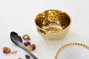 A slightly angled view of a Pampa Bay Golden Millennium Porcelain Snack Bowl. The bowl has a rich, hammered gold interior with a reflective, textured surface, and a smooth, matte off-white or cream exterior. The rim of the bowl is irregularly shaped, following the hammered texture of the interior. It is presented against a plain white background.