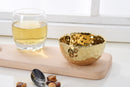 A slightly angled view of a Pampa Bay Golden Millennium Porcelain Snack Bowl. The bowl has a rich, hammered gold interior with a reflective, textured surface, and a smooth, matte off-white or cream exterior. The rim of the bowl is irregularly shaped, following the hammered texture of the interior. It is presented against a plain white background.