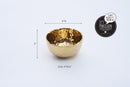 A slightly angled view of a Pampa Bay Golden Millennium Porcelain Snack Bowl. The bowl has a rich, hammered gold interior with a reflective, textured surface, and a smooth, matte off-white or cream exterior. The rim of the bowl is irregularly shaped, following the hammered texture of the interior. It is presented against a plain white background.