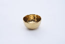 A slightly angled view of a Pampa Bay Golden Millennium Porcelain Snack Bowl. The bowl has a rich, hammered gold interior with a reflective, textured surface, and a smooth, matte off-white or cream exterior. The rim of the bowl is irregularly shaped, following the hammered texture of the interior. It is presented against a plain white background.